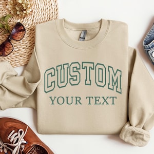 Puede incluir: Una sudadera de cuello redondo en color beige con el texto verde oscuro CUSTOM en una fuente universitaria arqueada, con las palabras YOUR TEXT impresas en línea recta debajo. La prenda tiene mangas largas y cuello, puños y dobladillo acanalados.