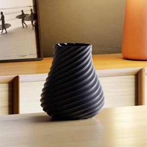 Pode incluir: Um vaso preto com um design em espiral. O vaso está sobre uma superfície de madeira. No fundo, uma fotografia a preto e branco emoldurada de pessoas com pranchas de surf.