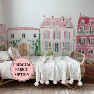Può includere: Una scena accogliente della camera da letto con una struttura del letto intrecciata e una coperta bianca a maglia a trecce con pompon. Lo sfondo è un murale di edifici in stile europeo rosa e bianco. Il testo "PREMIUM FABRIC OPTION" è visualizzato su un'etichetta rosa.