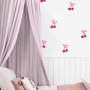 Könnte beinhalten: Ein rosa Himmelbett mit einem weißen Kopfteil und einer rosa-weißen Bettdecke. Die Wand hinter dem Bett ist mit roten Kirschen mit rosa Schleifen verziert.