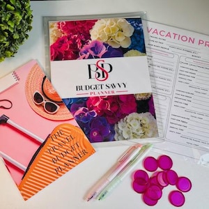 May include: Two pink and white planners with floral designs and the text "Budget Savvy Planner" and "The Travel Budget Planner". The planners are on a white surface with two pens and pink circle stickers.