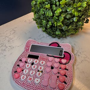 May include: A pink calculator with a Hello Kitty design, encrusted with rhinestones. The calculator has heart-shaped buttons and a digital display. The design includes a pink bow and a white border. The calculator is on a white surface.