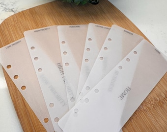 May include: A collection of translucent planner dividers with printed labels including "Low Priority," "High Priority," "Personal," "Saving Challenge," "Business," and "Home." The dividers have pre-punched holes for binding and are arranged on a wooden surface.