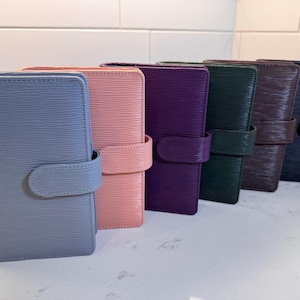 Puede incluir: Una colección de seis organizadores de cuero en varios colores, incluyendo azul, rosa, morado, verde, marrón y azul marino. Cada organizador presenta una superficie texturizada y un cierre de correa a juego.