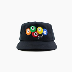 Peut inclure: Une casquette de baseball noire ornée d'une broderie représentant cinq boules de billard colorées disposées en groupe. Chaque boule affiche une lettre formant le mot LUCKY, avec le texte blanc en cursive you! brodé juste en dessous.