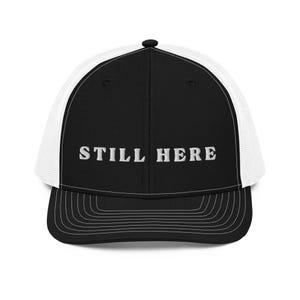 May include: Black and white trucker cap with the embroidered text "STILL HERE" in white. The hat has a black front panel and peak, with a white mesh back. A classic, casual accessory.