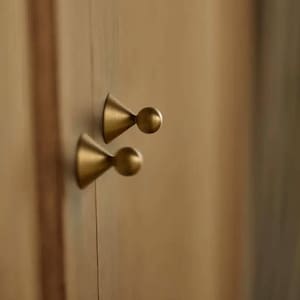 Puede incluir: Primer plano de dos tiradores de armario de latón. Cada tirador presenta una base cónica y un mango esférico. Los tiradores están montados en una puerta de armario de madera marrón claro, creando una estética minimalista y elegante.
