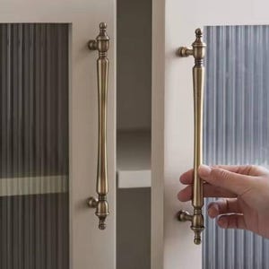 May include: Cream-colored cabinet doors with ribbed glass panels and antique brass handles. One door is slightly ajar, revealing a glimpse of the interior. The handles are long and ornate, adding a touch of vintage charm to the furniture.