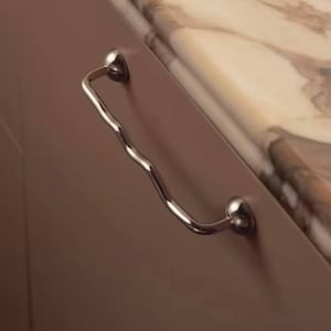 May include: A chrome cabinet pull with a wavy handle design. The pull is mounted on a taupe cabinet door, with a marble countertop in the background. The pull has a modern, minimalist aesthetic.