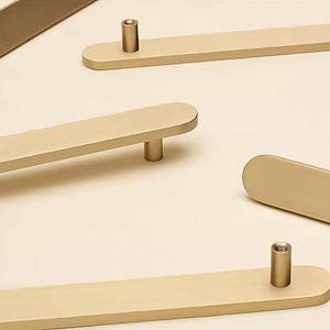 May include: Close-up of several gold-colored cabinet pulls. Each pull has a rectangular bar with rounded ends and a cylindrical post for mounting. The hardware is a warm, neutral tone, suitable for various cabinet styles.