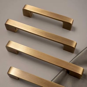 Puede incluir: Cuatro tiradores de armario en tono lat&oacute;n dispuestos sobre una superficie gris. Cada tirador tiene una barra rectangular con bordes curvos y un acabado ligeramente texturizado. Dise&ntilde;ados para un agarre f&aacute;cil, a&ntilde;aden elegancia. (Medidas en cm)