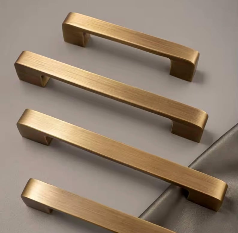 Puede incluir: Cuatro tiradores de armario en tono lat&oacute;n dispuestos sobre una superficie gris. Cada tirador tiene una barra rectangular con bordes curvos y un acabado ligeramente texturizado. Dise&ntilde;ados para un agarre f&aacute;cil, a&ntilde;aden elegancia. (Medidas en cm)