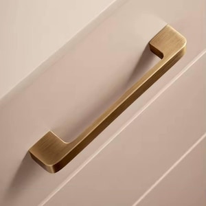Puede incluir: Un tirador de armario dorado con un dise&ntilde;o rectangular. El tirador est&aacute; montado en una puerta de armario rosa claro. El tirador es de metal y tiene un acabado cepillado.