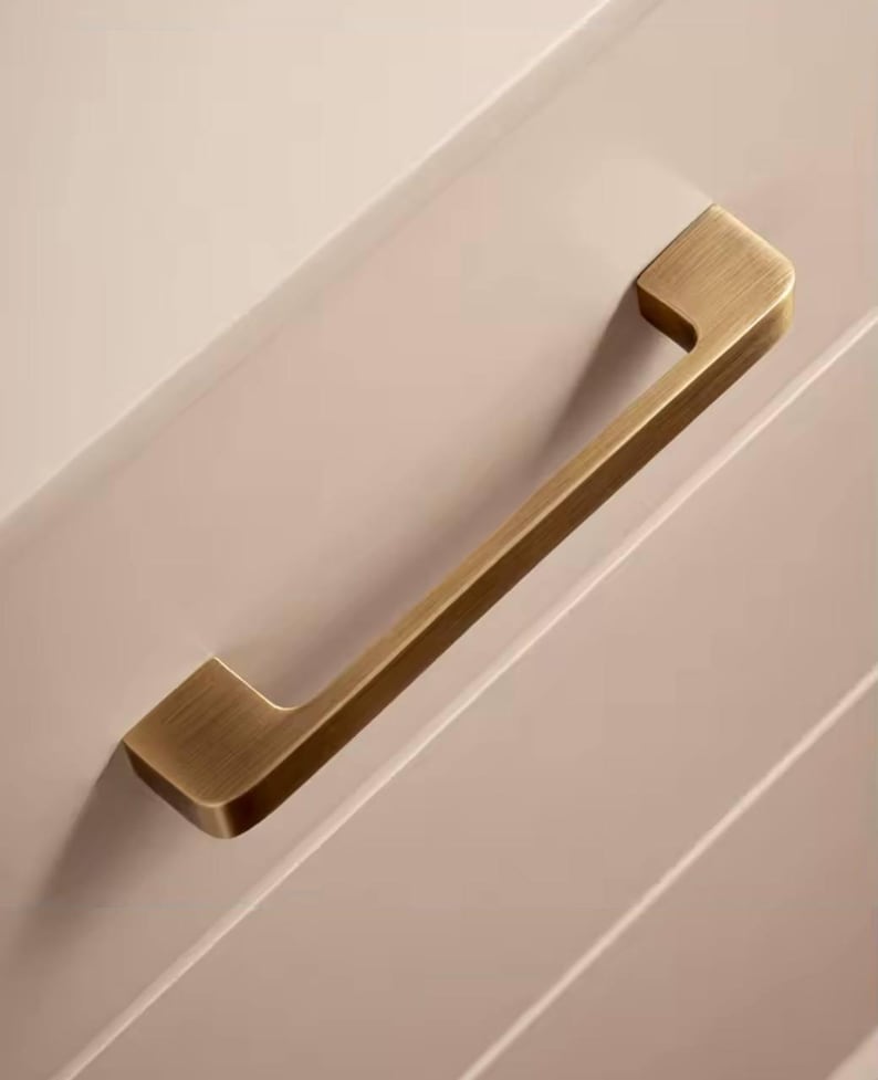 Puede incluir: Un tirador de armario dorado con un dise&ntilde;o rectangular. El tirador est&aacute; montado en una puerta de armario rosa claro. El tirador es de metal y tiene un acabado cepillado.