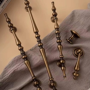 May include: An assortment of antique bronze cabinet hardware, including long pulls, a knob, and a smaller pull. The hardware features ornate detailing and a warm, aged finish, suggesting a vintage or classic aesthetic. The pieces are arranged on a neutral fabric.