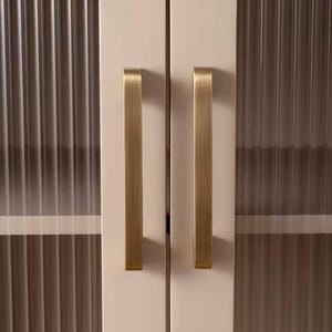 Puede incluir: Primer plano de una puerta de armario con un panel de vidrio acanalado y un tirador dorado cepillado. La puerta del armario es de color crema claro. El tirador es rectangular y est&aacute; orientado verticalmente.
