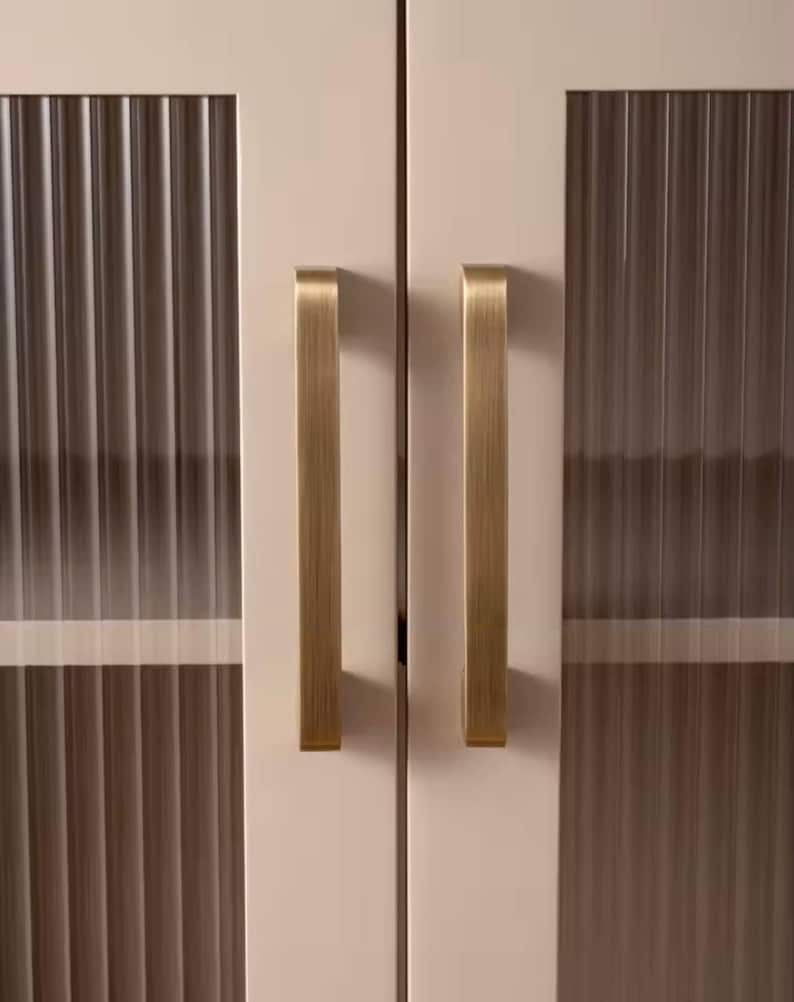 Puede incluir: Primer plano de una puerta de armario con un panel de vidrio acanalado y un tirador dorado cepillado. La puerta del armario es de color crema claro. El tirador es rectangular y est&aacute; orientado verticalmente.
