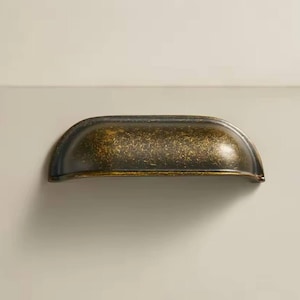 May include: A bronze-colored, half-moon shaped drawer pull. The hardware has a textured surface with a speckled appearance. The drawer pull is mounted on a light beige surface. This hardware is suitable for cabinets and drawers.