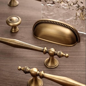 May include: An assortment of antique brass cabinet hardware, including drawer pulls, knobs, and a cup pull. The hardware features ornate detailing and a warm, aged finish, perfect for vintage-style furniture.