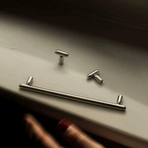 Puede incluir: Un juego de herrajes plateados para armarios, que incluye un tirador largo y dos pomos en forma de T. Los herrajes se muestran sobre una superficie de color claro, lo que sugiere una estética de diseño moderno o minimalista.