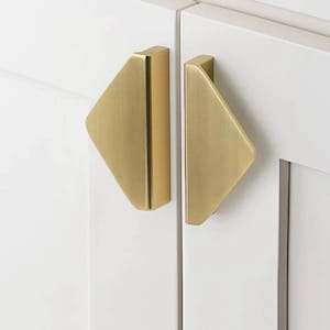 May include: Two gold-coloured cabinet pulls with a geometric design. Each pull has a triangular shape with a rectangular handle. The pulls are mounted on a white cabinet door.