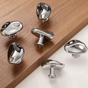 Może przedstawiać: Sześć polerowanych chromowanych gałek do szafek o organicznym, przypominającym kamień kształcie. Gałki mają gładką, odblaskową powierzchnię i są zamontowane na krótkich, cylindrycznych trzpieniach. Ułożone na jasnobrązowej drewnianej powierzchni.