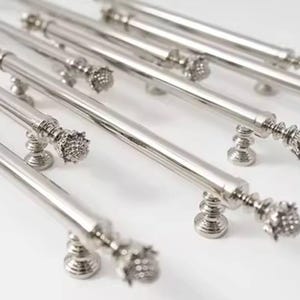 Peut inclure: Plusieurs poignées de meubles argentées polies, avec des extrémités décoratives en forme de fleur, sont disposées sur une surface blanche. Les poignées ont une barre cylindrique.