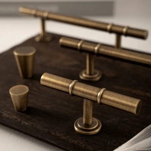 Op de afbeelding: Een verzameling antieke messing kastbeslagen, waaronder cilindrische stanggrepen en conische knoppen. De hardware wordt gepresenteerd op een donker houten oppervlak, waarbij de verouderde afwerking en het ontwerp worden benadrukt. De grepen zijn ongeveer 15 cm lang.