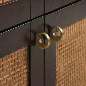 May include: Close-up of a cabinet door with a woven rattan texture and dark brown trim. The cabinet features two brass-colored ring pulls. The design combines natural materials with a modern aesthetic, suitable for home decor.