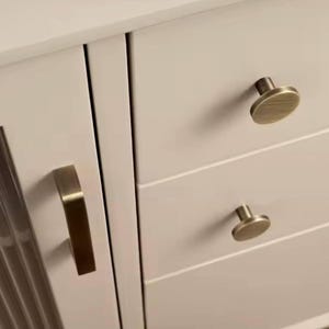 Puede incluir: Primer plano de un armario color crema con herrajes de lat&oacute;n. El armario presenta un tirador vertical en la puerta y pomos redondos en los cajones. El armario tiene un dise&ntilde;o moderno.