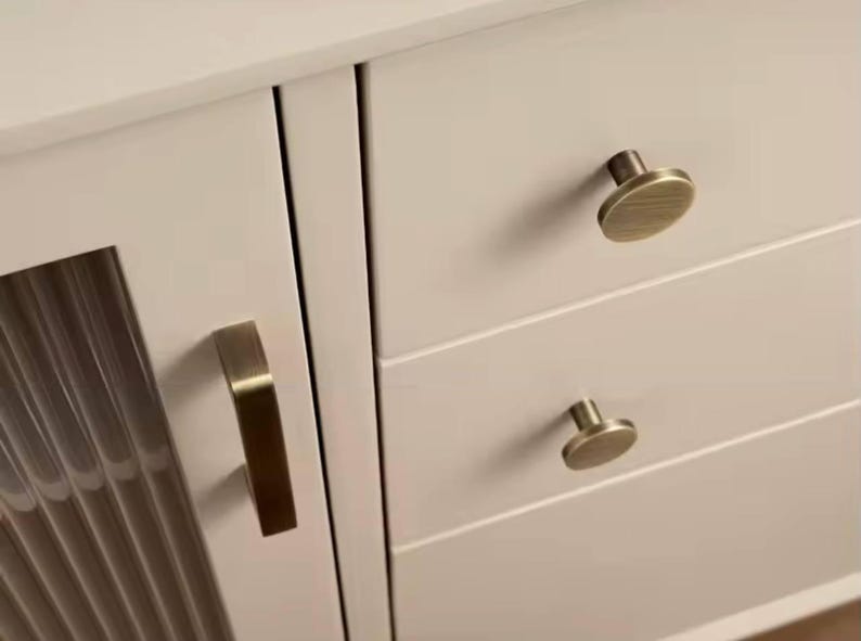 Puede incluir: Primer plano de un armario color crema con herrajes de lat&oacute;n. El armario presenta un tirador vertical en la puerta y pomos redondos en los cajones. El armario tiene un dise&ntilde;o moderno.