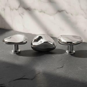 May include: Three polished chrome cabinet knobs with organic, pebble-like shapes. The knobs have a reflective surface and are mounted on short, cylindrical stems. The knobs are arranged on a dark, textured surface against a marble background.
