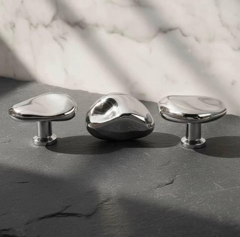 May include: Three polished chrome cabinet knobs with organic, pebble-like shapes. The knobs have a reflective surface and are mounted on short, cylindrical stems. The knobs are arranged on a dark, textured surface against a marble background.