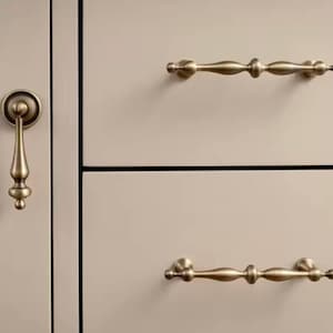 Puede incluir: Primer plano de herrajes de gabinete con manijas y un tirador de latón antiguo. Las manijas son horizontales, con un diseño decorativo y alargado. El tirador es vertical, con forma de lágrima. Las puertas son de color beige neutro.