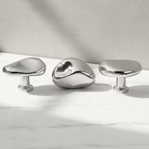 May include: Three polished chrome cabinet knobs with organic, pebble-like shapes. The knobs have a smooth, reflective surface and are mounted on short, cylindrical stems. The design is modern and minimalist.