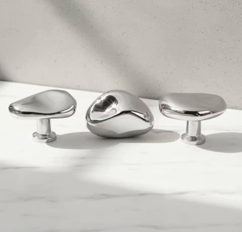 May include: Three polished chrome cabinet knobs with organic, pebble-like shapes. The knobs have a smooth, reflective surface and are mounted on short, cylindrical stems. The design is modern and minimalist.