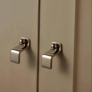 Op de afbeelding: Twee grepen voor kasten van geborsteld nikkel zijn gemonteerd op een lichtbeige kastdeur. Elke greep heeft een kleine, ronde knop boven een gebogen, rechthoekige handgreep. De hardware is ontworpen voor een moderne esthetiek.