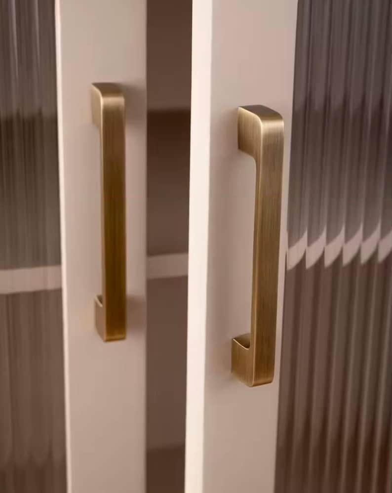 Puede incluir: Primer plano de una puerta de armario blanca con un tirador vertical de color lat&oacute;n antiguo. La puerta tiene un panel de vidrio texturizado y acanalado. El tirador es rectangular con un agarre curvado.