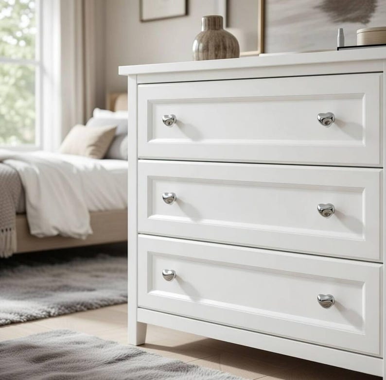 May include: A white three-drawer dresser with silver-toned knobs. The dresser has a classic design with recessed panels on each drawer. A decorative vase sits on top of the dresser. The bedroom setting includes a bed and a window.
