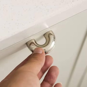 Op de afbeelding: Een gepolijste, verchroomde kastgreep in U-vorm. De greep is gemonteerd op een witte kastdeur. De afbeelding toont een hand die de greep vastgrijpt, wat suggereert dat deze wordt gebruikt om een kast of lade te openen.