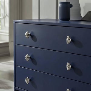 May include: A navy blue dresser with four drawers and silver-toned knobs. A small, dark blue ceramic vase sits on top of the dresser. The dresser is a modern style and is in a well-lit room.