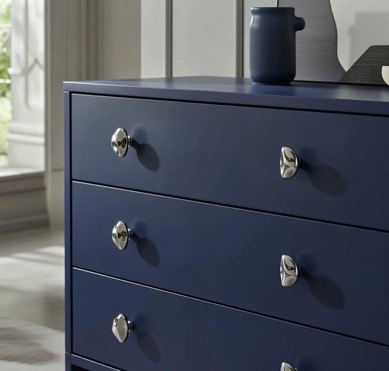 May include: A navy blue dresser with four drawers and silver-toned knobs. A small, dark blue ceramic vase sits on top of the dresser. The dresser is a modern style and is in a well-lit room.