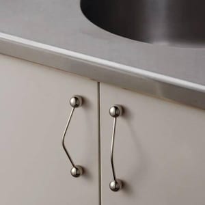 May include: Close-up of a kitchen cabinet with two doors and brushed metal hardware. The cabinet doors are a light neutral color. The hardware is a curved design with a ball at each end.