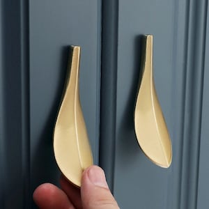 May include: Two gold-coloured cabinet pulls with a leaf-like design. The pulls are mounted on a blue-grey cabinet door. The pulls have a smooth, polished finish and are slightly curved.