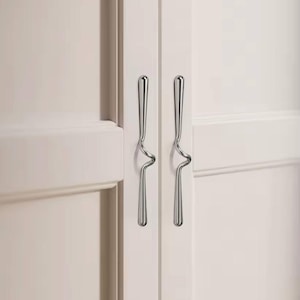 Pode incluir: Close-up de duas portas de armário com puxadores prateados polidos. Os puxadores têm um design curvo único, adicionando um toque moderno às portas brancas do armário. Os puxadores estão orientados verticalmente.
