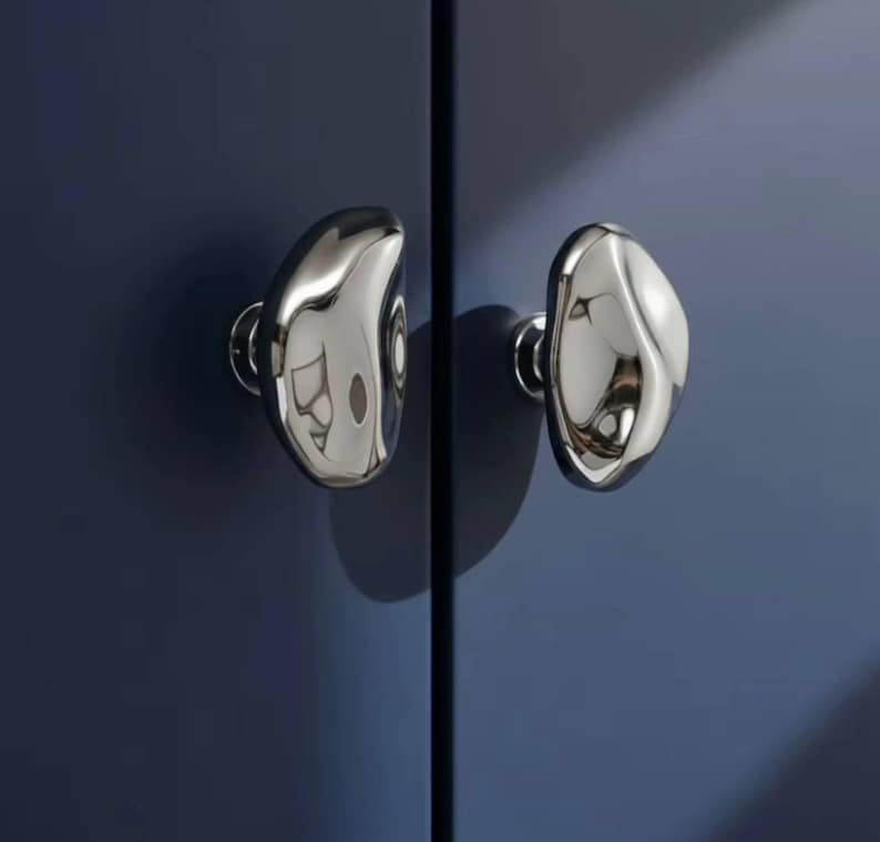 May include: Two polished chrome cabinet knobs with an organic, pebble-like shape are mounted on a dark blue cabinet door. The knobs reflect the surrounding environment, creating a mirrored effect. The hardware adds a modern touch to the cabinet.