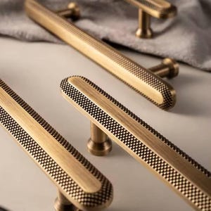 Op de afbeelding: Close-up van verschillende antieke messing kastgrepen. De grepen hebben een rechthoekige vorm met een getextureerd, geblokt patroon aan de zijkanten en een glad middenpaneel. De hardware is ontworpen voor meubels en woondecoratie.