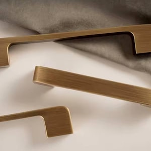Puede incluir: Tres tiradores de armario de color bronce con un dise&ntilde;o rectangular. Los tiradores est&aacute;n dispuestos sobre una superficie blanca con una tela gris en el fondo. El herraje tiene un acabado cepillado.