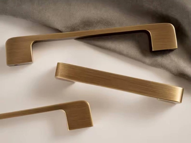 Puede incluir: Tres tiradores de armario de color bronce con un dise&ntilde;o rectangular. Los tiradores est&aacute;n dispuestos sobre una superficie blanca con una tela gris en el fondo. El herraje tiene un acabado cepillado.