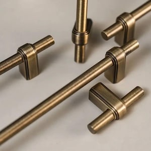 Puede incluir: Primer plano de varios tiradores y pomos de armario de latón antiguo. El herraje presenta un diseño en forma de T con un mango cilíndrico y una base cuadrada. El acabado tiene un aspecto cálido y envejecido, lo que sugiere una estética vintage. Los pomos y tiradores están dispuestos sobre una superficie neutra.